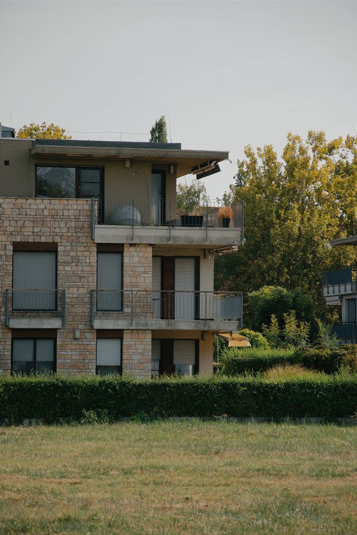 contact-img Modern Hungarian apartment building with stone facade and lush greenery on a summer day.